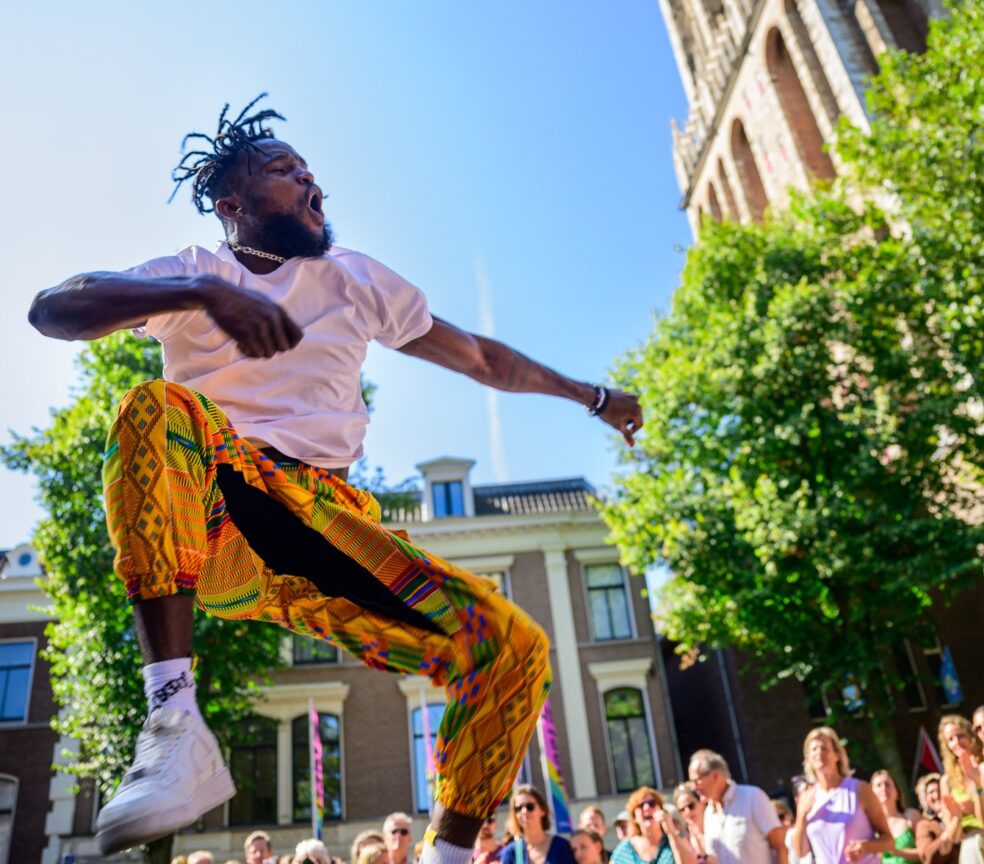 Foto van UITFeest 2024, een man met vlechtjes, in wit t-shirt en felgekleurde broek, springt in de lucht, met één knie opgetrokken. Er staan mensen omheen te kijken, een klein stukje van de Dom staat op de foto.